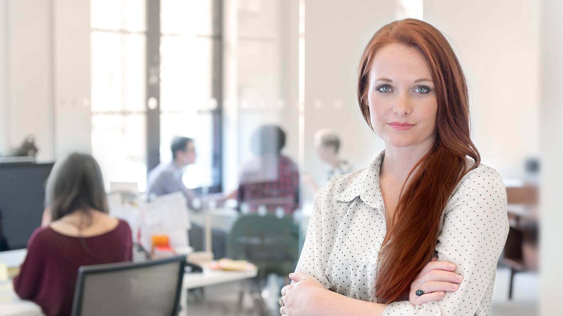 Eine Frau mit langen roten Haaren steht selbstbewusst mit verschränkten Armen in einem modernen Büro. Sie trägt eine weiße Bluse mit kleinen Punkten. Im Hintergrund sind unscharf weitere Personen an Schreibtischen zu sehen, die gemeinsam arbeiten.