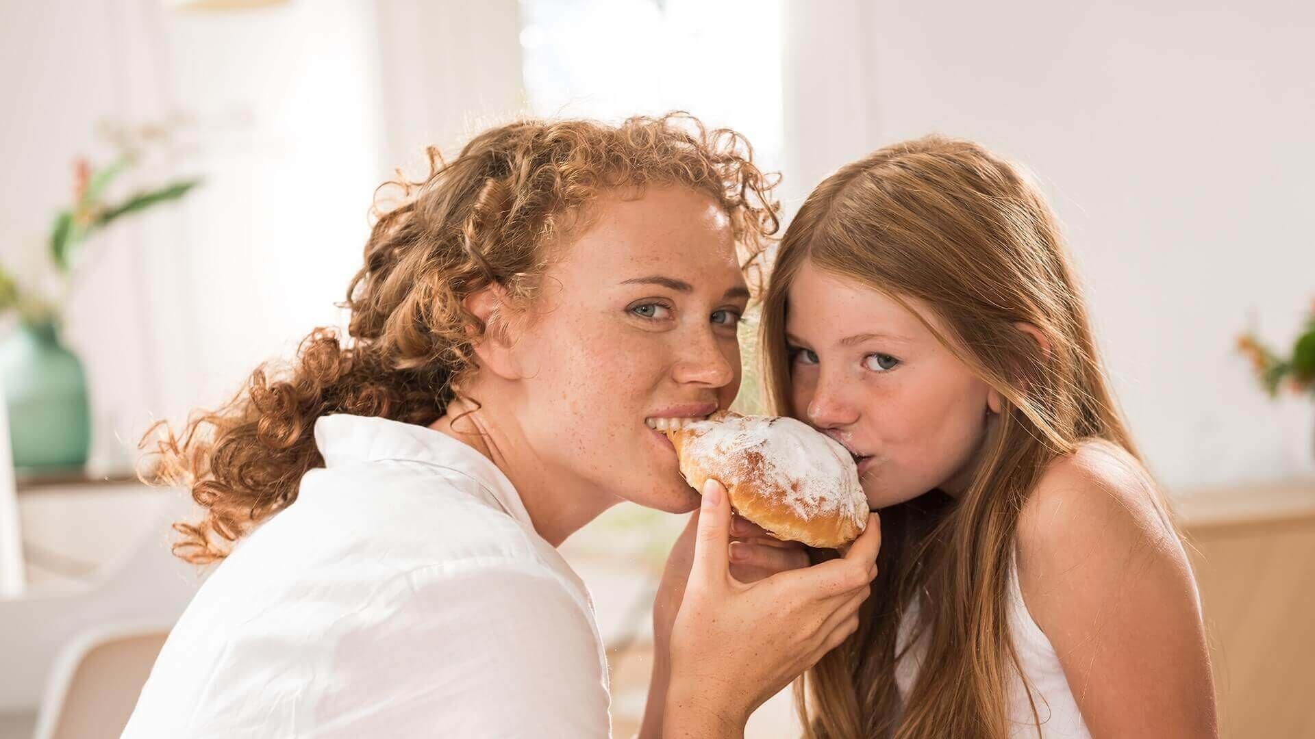 Eine Frau mit lockigen, roten Haaren und ein Mädchen mit langen, glatten, braunen Haaren teilen sich einen mit Puderzucker bestreuten Krapfen. Beide lächeln und beißen gleichzeitig in das Gebäck. Im Hintergrund ist ein heller Raum mit unscharfen Details zu sehen.